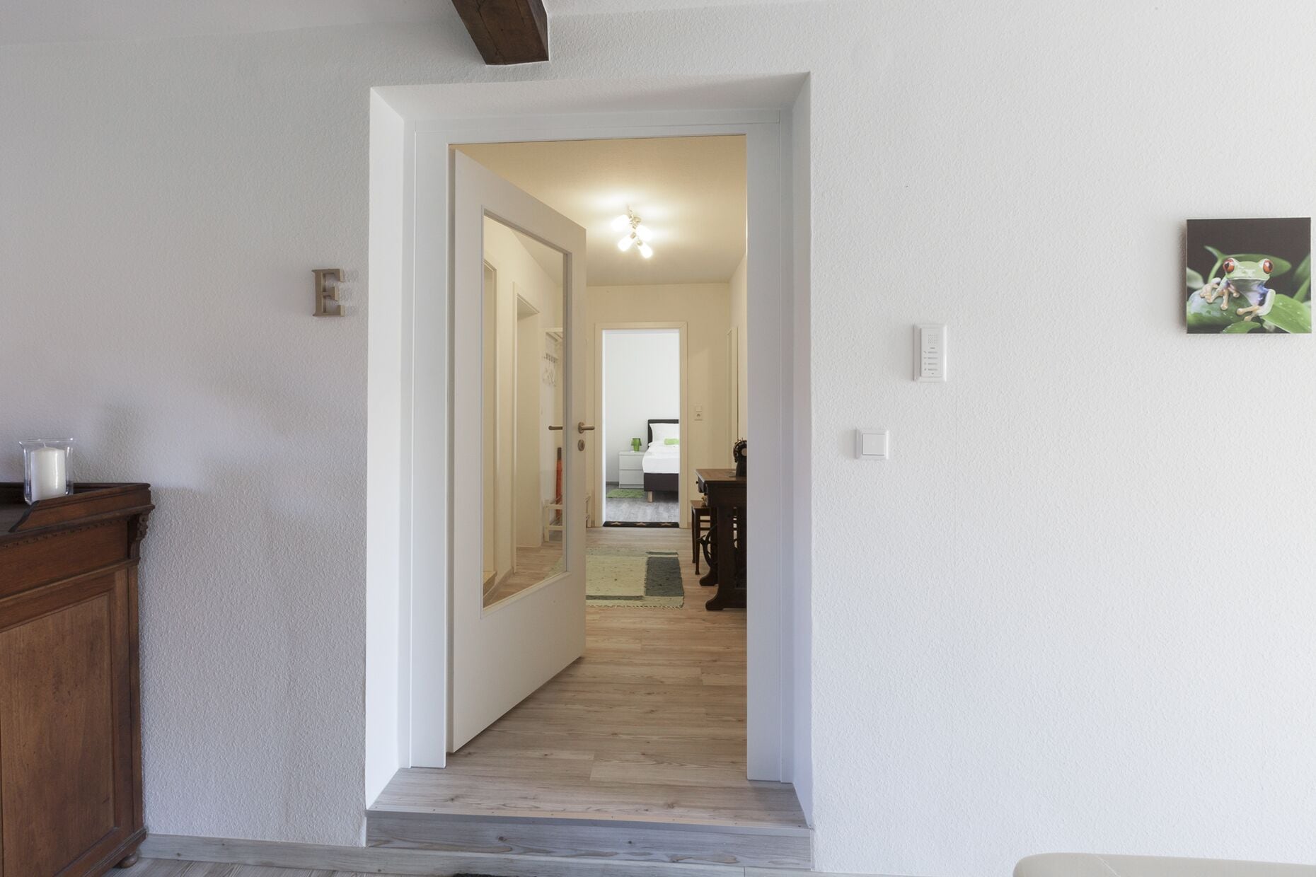 Corredor que leva a um quarto com uma cama, um criado-mudo e uma mesa de cabeceira. Há uma porta à esquerda e um candeeiro fixado na parede acima da porta.