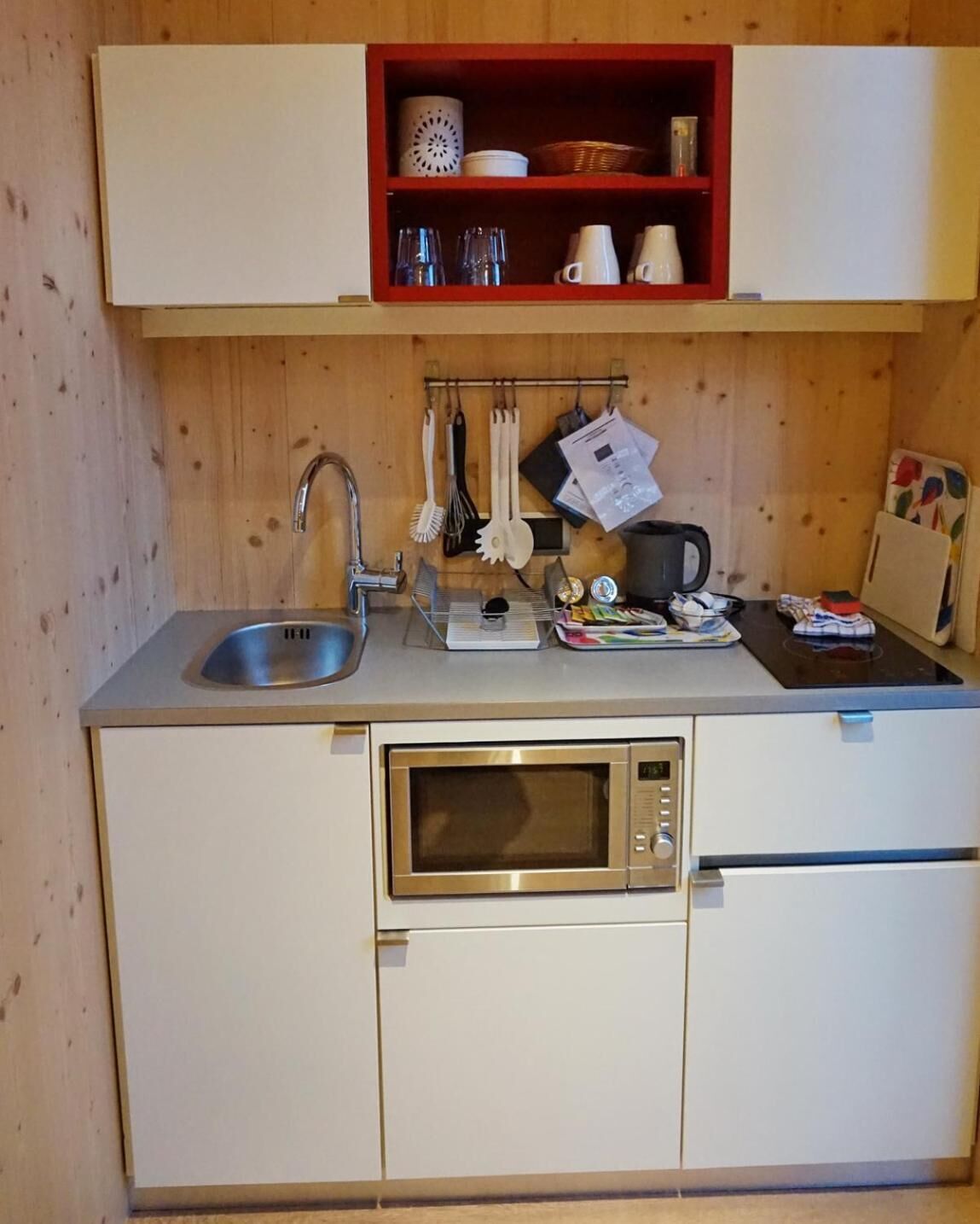 Una cucina compatta con un lavello in acciaio inossidabile, un forno a microonde e uno scaffale rosso sopra il bancone, con diversi oggetti da cucina.