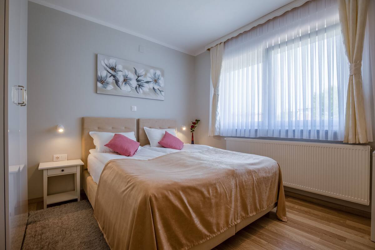 A bedroom with a large bed, a nightstand, a wall-mounted light, a radiator, and a window with sheer curtains.