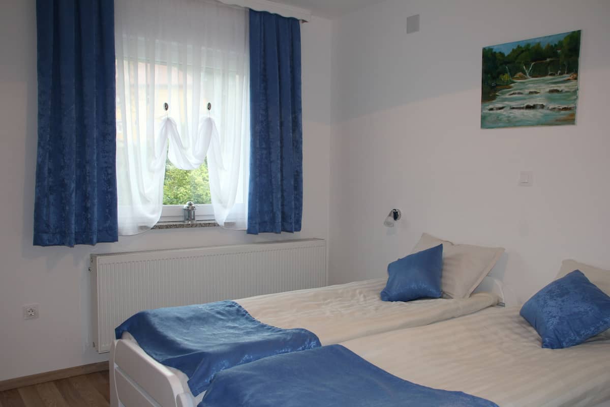 A bedroom with two beds, a window with blue curtains, a radiator, and a painting on the wall.