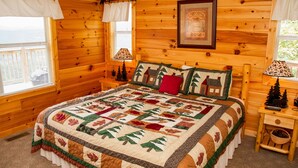 Un dormitori d'una cabana de fusta acollidora amb un llit gran, dues taules de nit, un quadre amb marc a la paret i una finestra amb cortines.