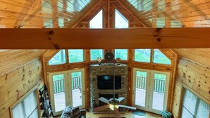 A cabin interior with a stone fireplace, wooden beams, and large windows offering views of the outdoors. - Paradise Found - Cabin (Sevierville)