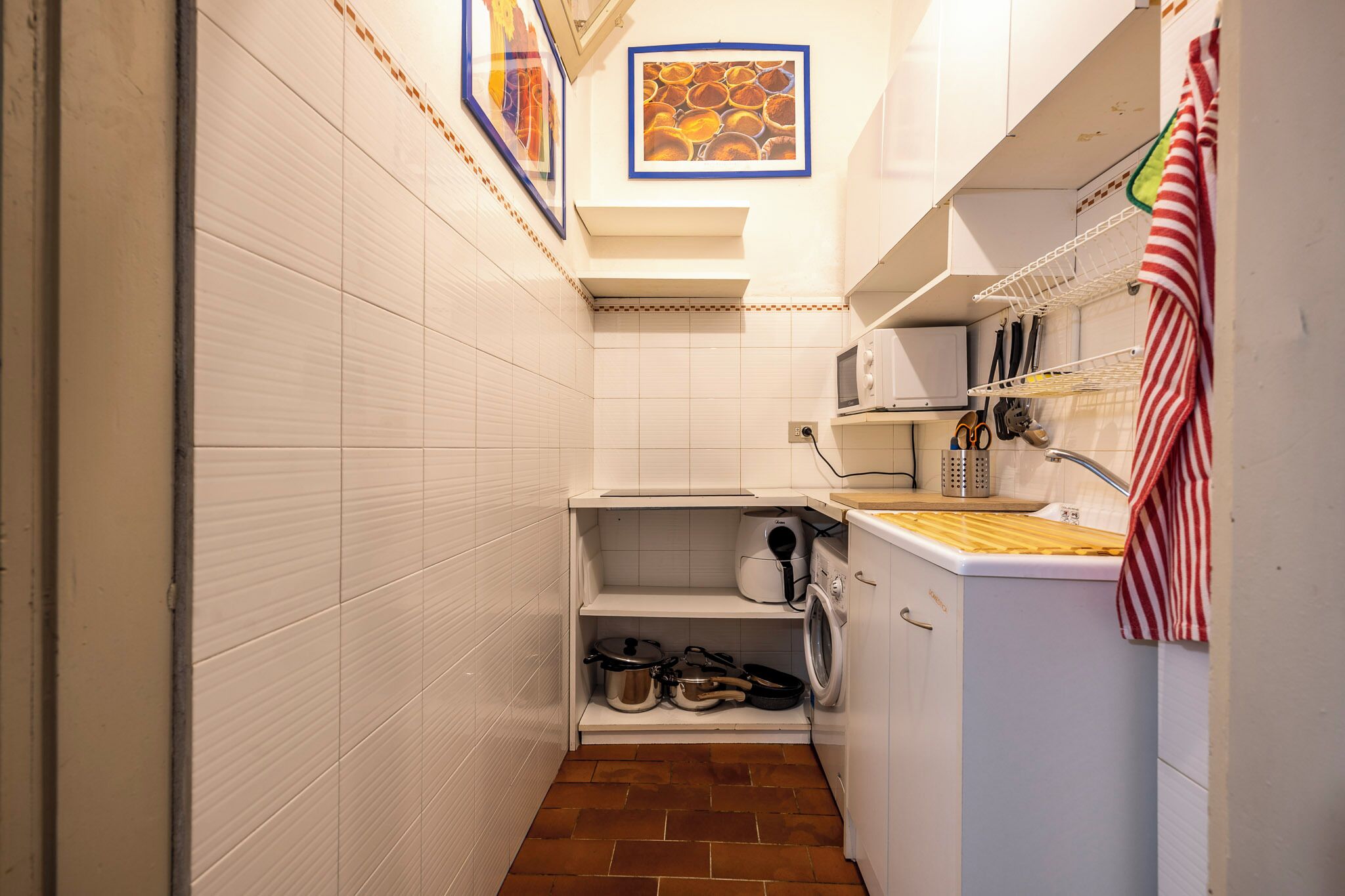 A compact kitchen with white tiled walls, a tiled floor, a microwave, a sink, and a small shelf with pots and pans.