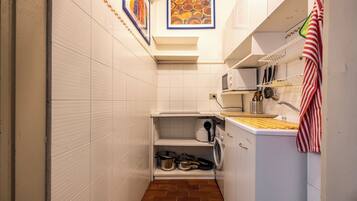 A compact kitchen with white tiled walls, a tiled floor, a microwave, a sink, and a small shelf with pots and pans.