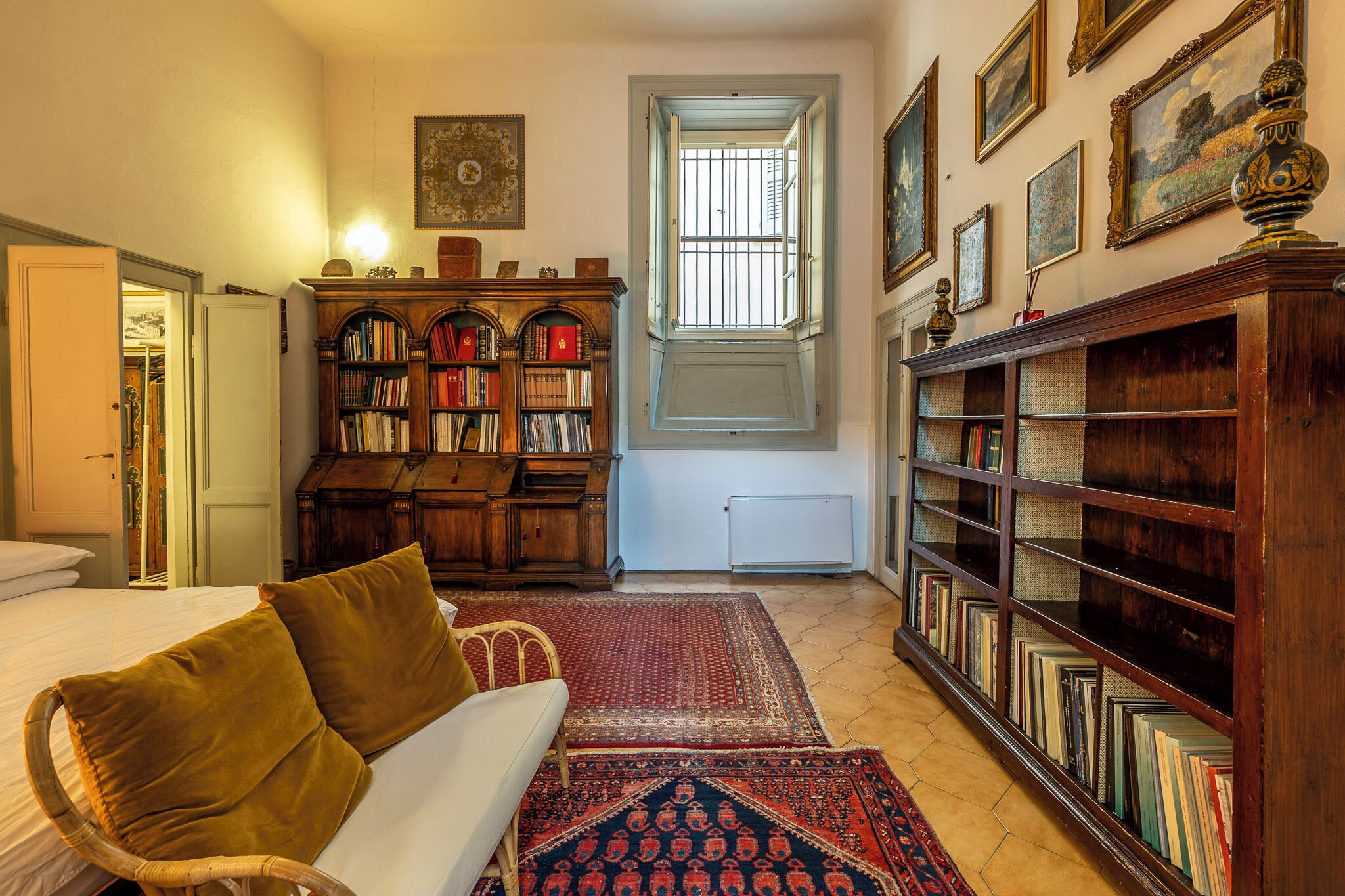 Une pièce avec un lit, une bibliothèque en bois, un tapis rouge à motifs et une fenêtre avec des barreaux.