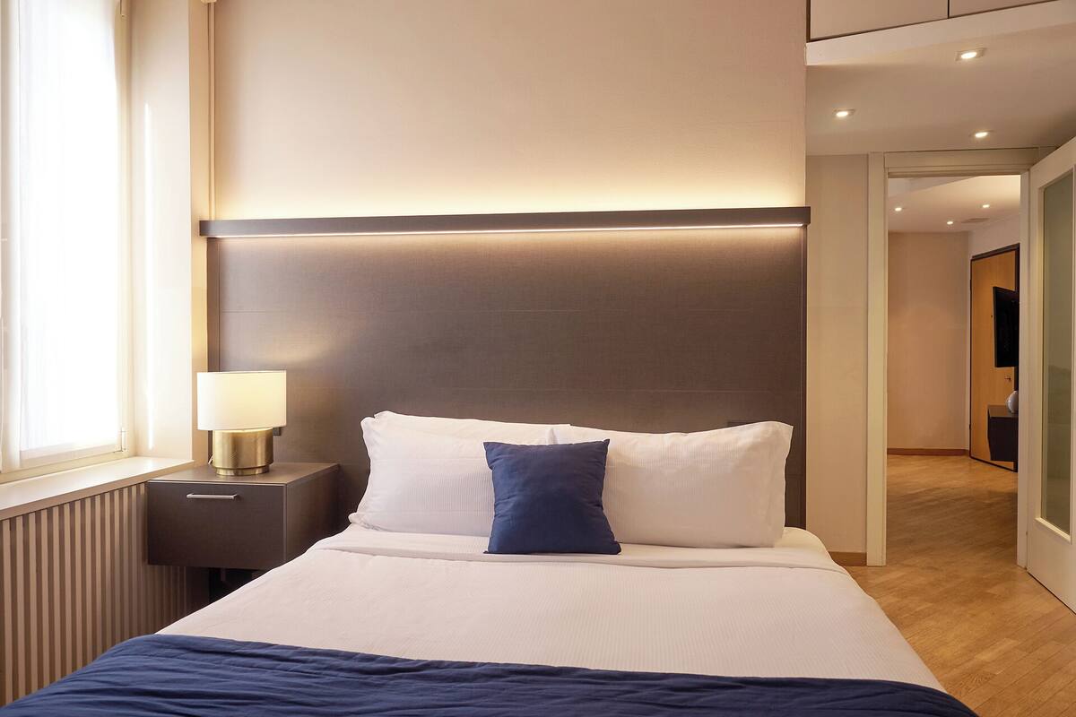 A modern hotel room with a large bed, a bedside lamp, and a view of the hallway.