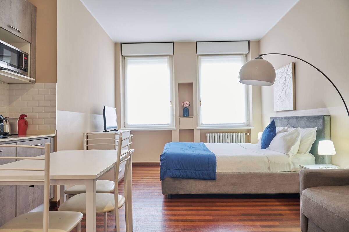 A compact hotel room with a bed, bedside table, lamp, and a kitchenette area.