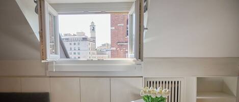 A room with a white table, a vase with flowers, a white chair, a grey couch, and a window with a view of buildings.