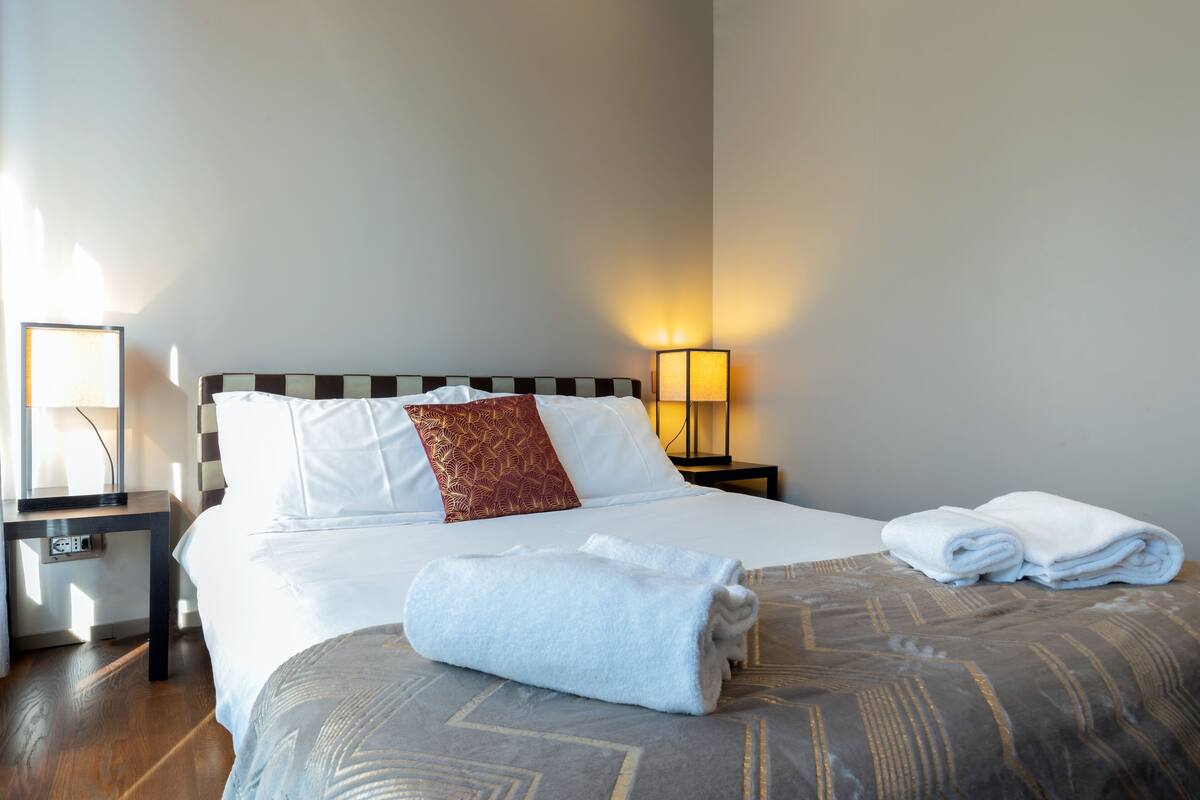 A neatly made bed with white linens and a patterned throw, a bedside lamp, and a folded towel on the bed.