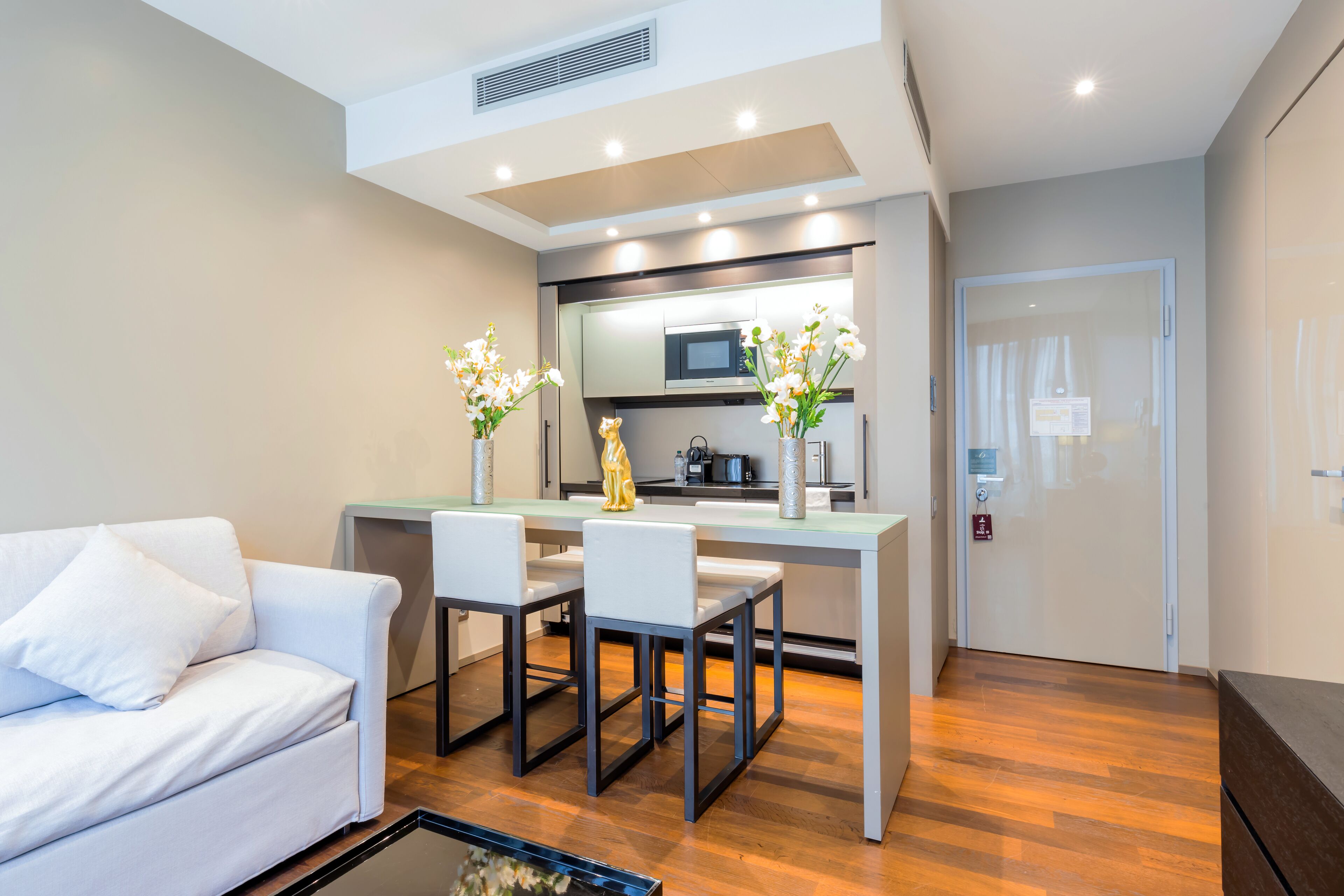 A modern living room with a white sofa, a dining table with chairs, a kitchen area with a microwave, and a door with a sign.