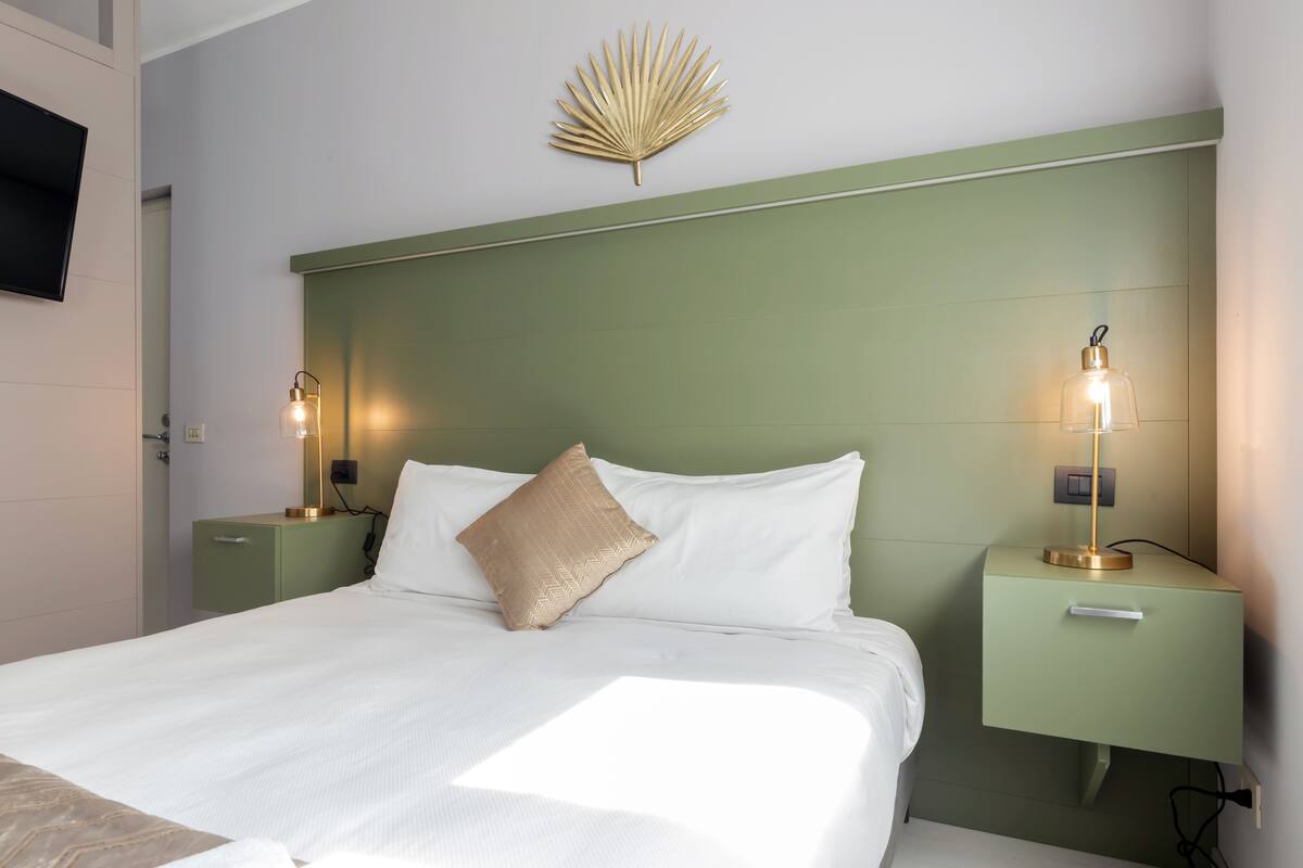 A neatly made bed with white linens and a beige pillow, flanked by two bedside tables with lamps, in a room with a green headboard and a wall-mounted fan.
