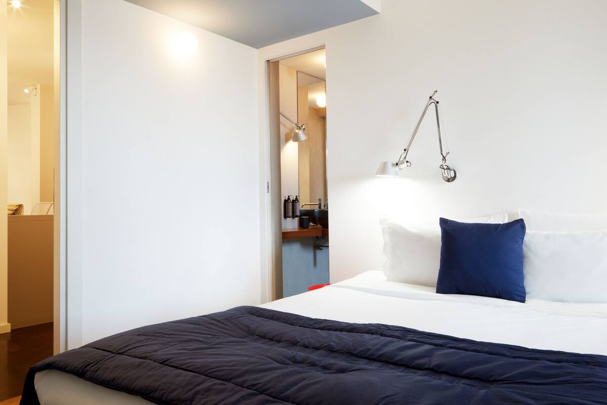 A modern hotel room with a neatly made bed, a large mirror, and a hanging lamp.