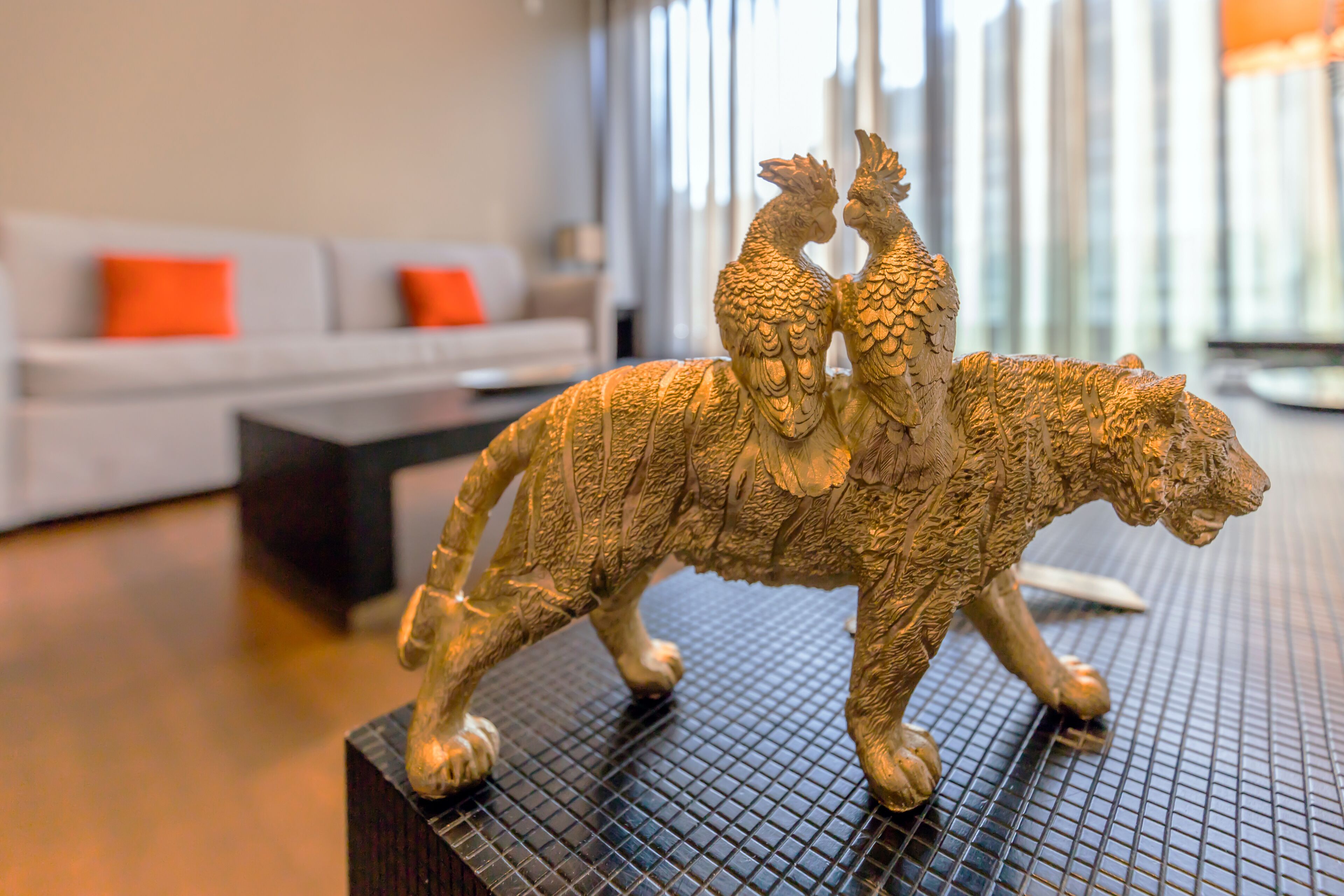 Una estatua de león dorada sobre un pedestal negro texturizado en un salón moderno con un sofá gris y cojines anaranjados.