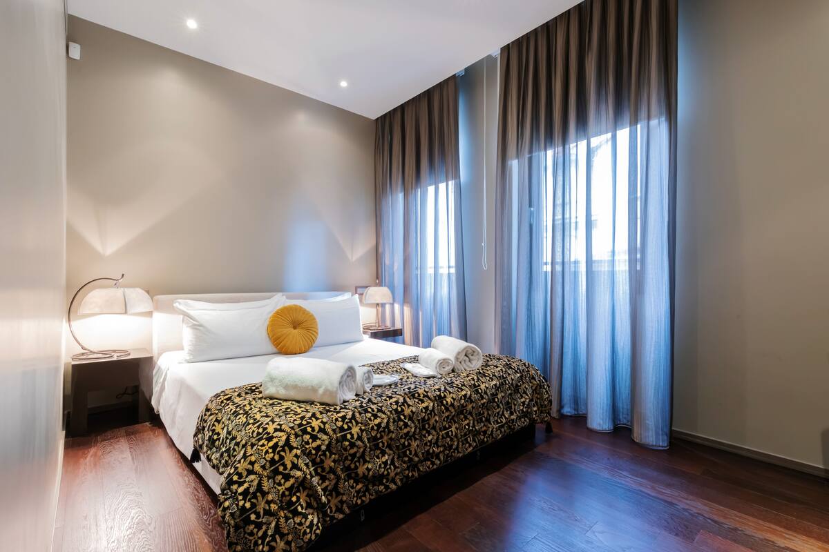 A hotel room with a large bed, a bedside table with a lamp, and a view of the city through sheer curtains.