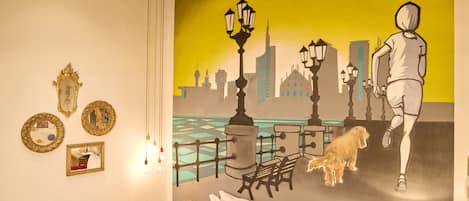 A hotel room with a bed, a red chair, a small table with a lamp, and a wall mural of a cityscape with a dog and a person running.