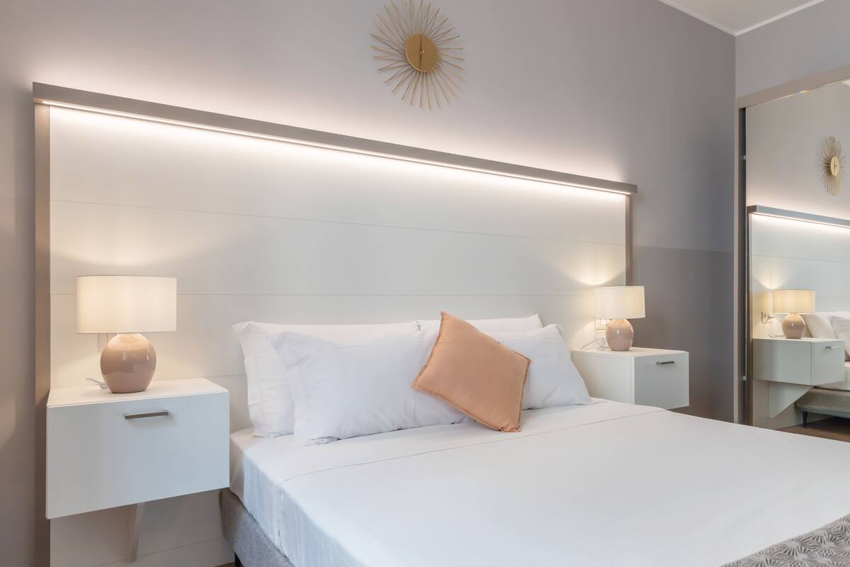 A modern hotel room with a large bed, white bedding, two bedside tables with lamps, and a large mirror on the wall.