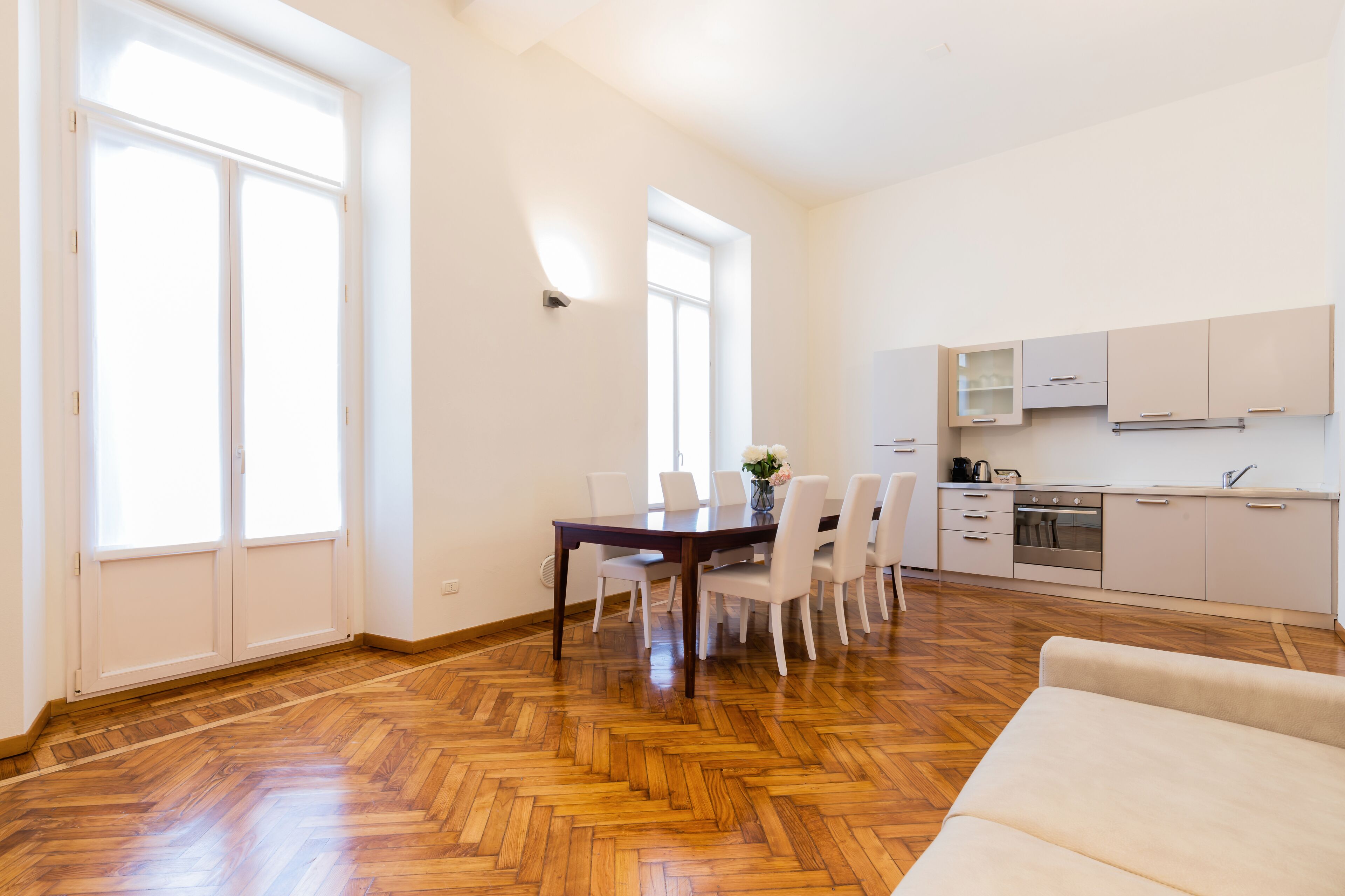 A spacious living area with a wooden herringbone floor, a dining table with chairs, a sofa, and a kitchen with cabinets and appliances.