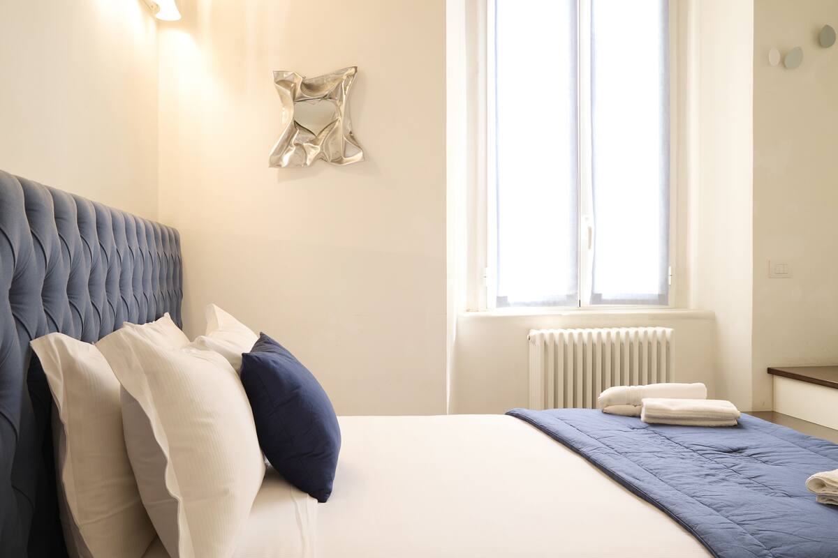 A neatly made bed with pillows and a blue blanket, a radiator, and a window with curtains.