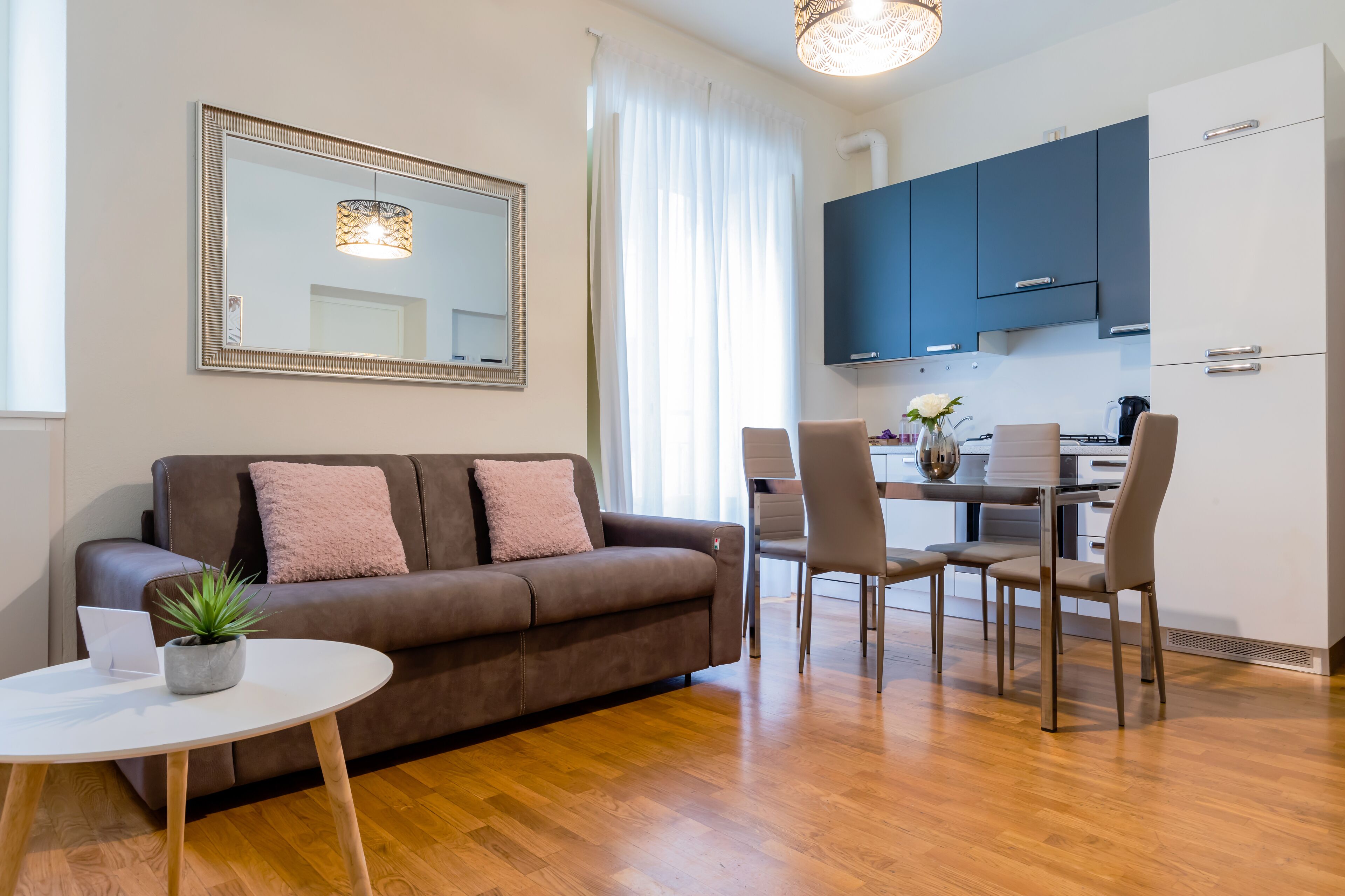 Una sala de estar moderna con un sofá marrón, una mesa redonda blanca y una pequeña planta en maceta. Un espejo grande cuelga en la pared, y se ve un comedor con sillas y una mesa al fondo.