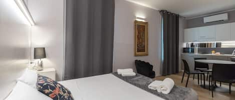 A modern hotel room with a neatly made bed, a small desk, and a kitchenette.