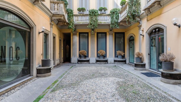 Un cortile con pavimento in pietra, piante in vaso e facciata di un edificio con ampie finestre e un portone.
