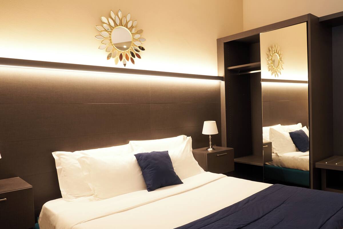 A neatly made bed with white linens and a dark blue pillow, a bedside table with a lamp, and a wall-mounted mirror with a sun-like design.