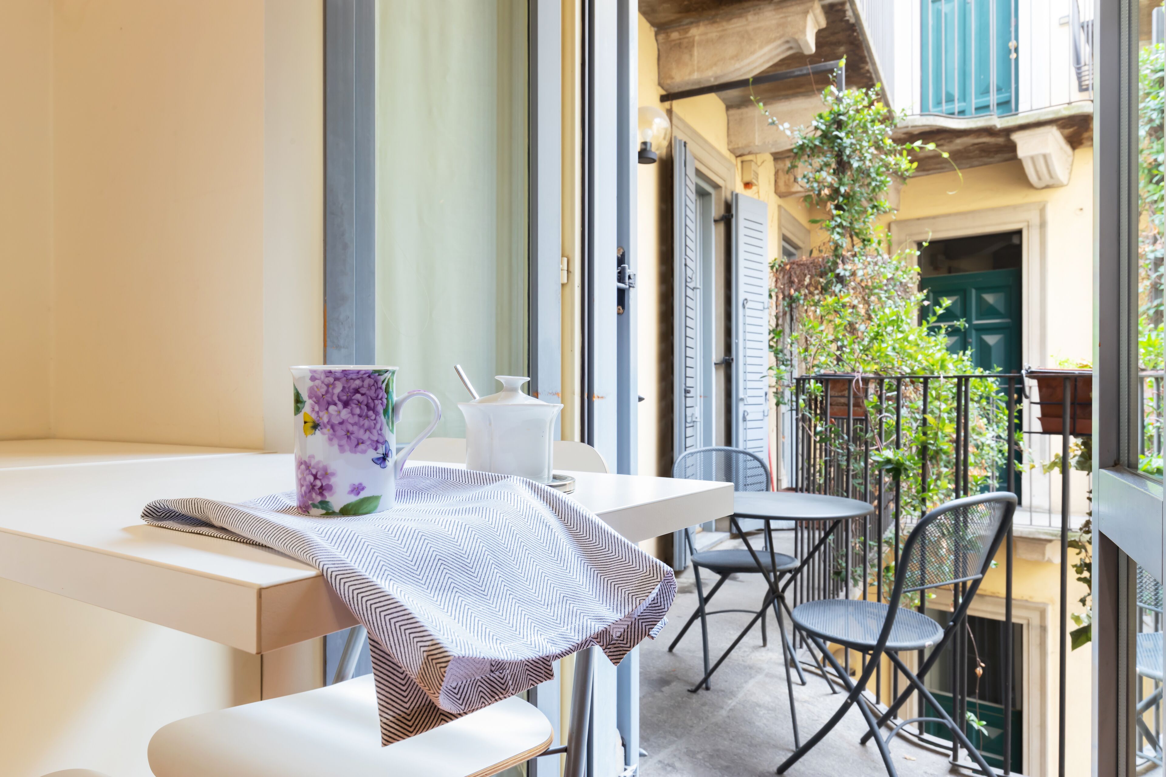 A balcony with a table and chairs, a potted plant, and a view of a building with balconies.