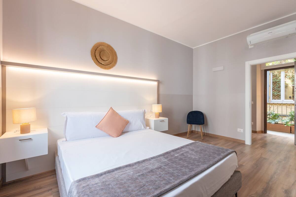 A modern bedroom with a large bed, bedside tables with lamps, a round wall decoration, and a view of the outdoors.
