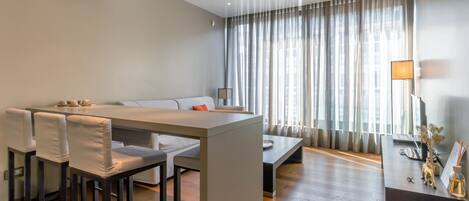 A modern hotel room with a dining area, a sofa, a coffee table, and a large window with curtains.