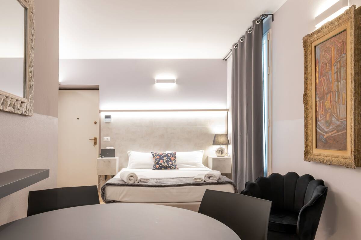 A hotel room with a bed, a desk, a chair, a lamp, and a painting on the wall.