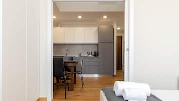 A modern hotel room with a wooden floor, a bed with a grey headboard, and a small kitchenette in the background.