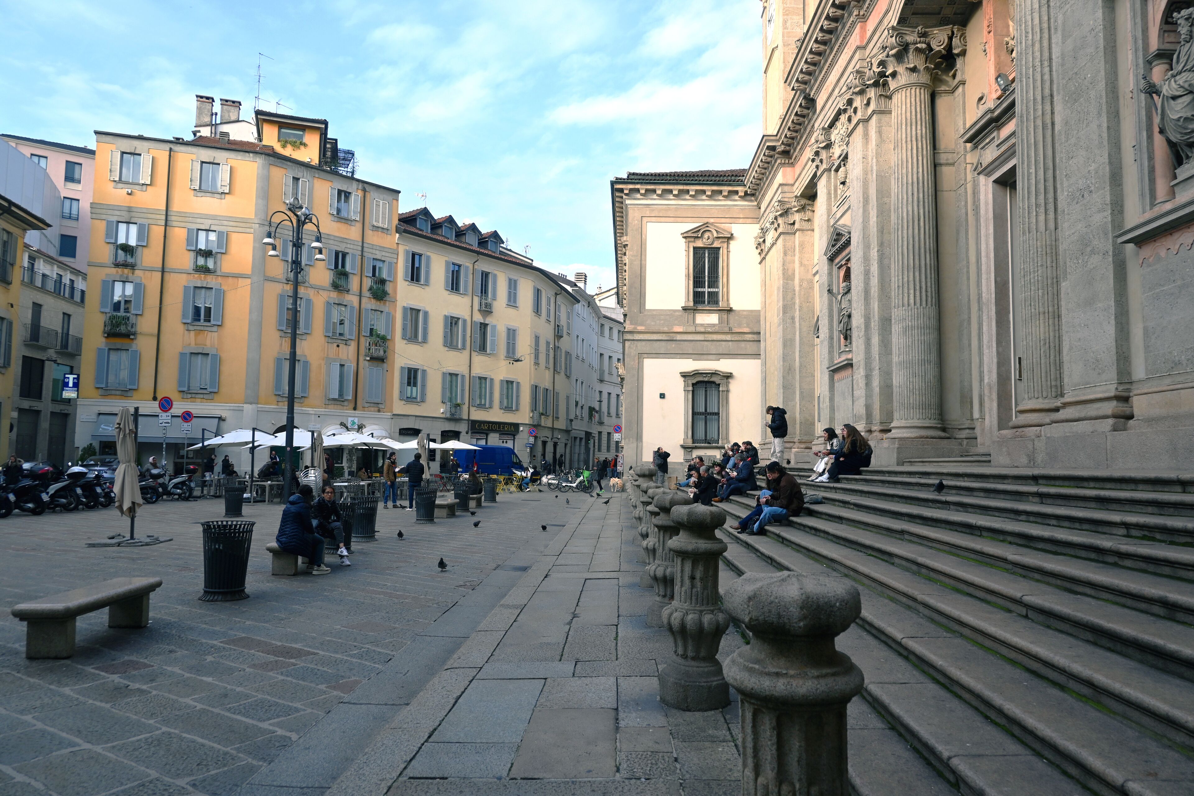 A public square with stone benches, a fountain, and classical architecture.