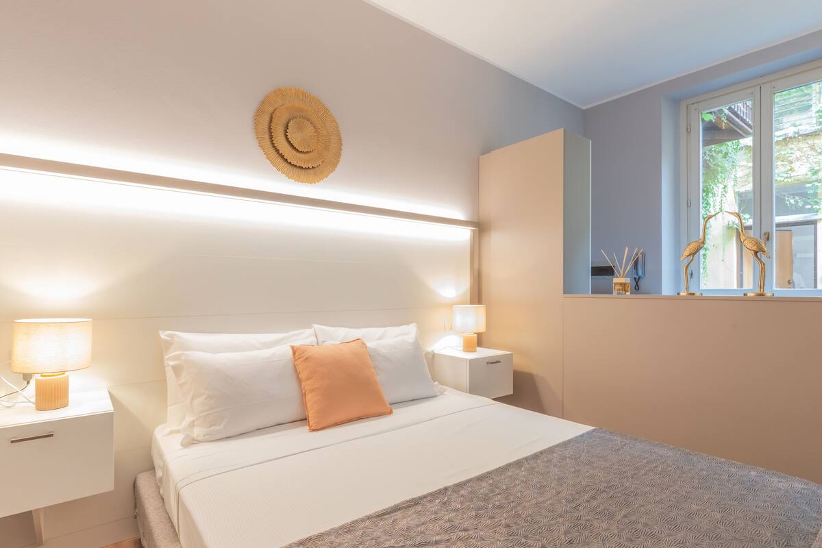 A neatly made bed with white and orange pillows, a patterned throw, and bedside lamps.