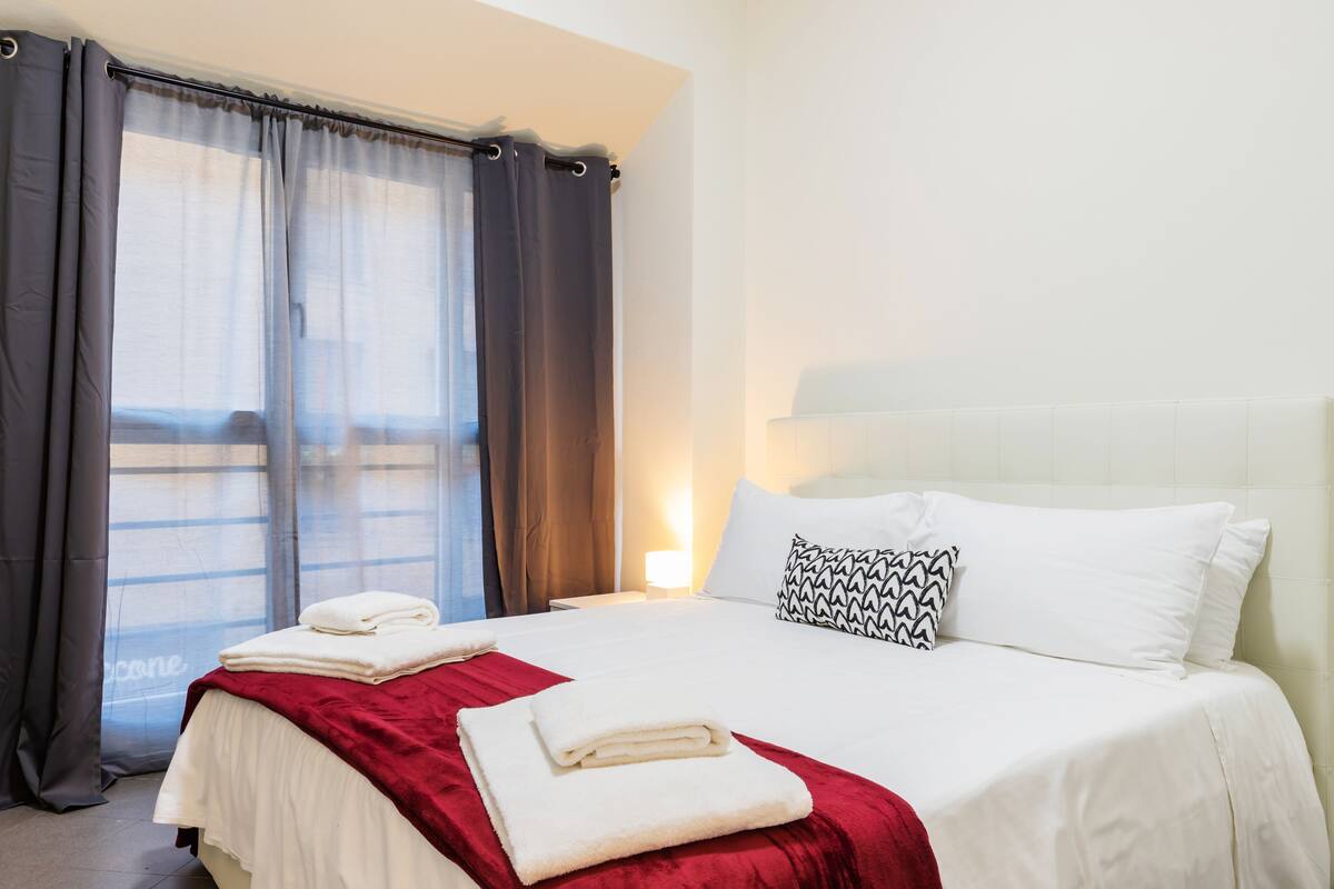 A hotel room with a neatly made bed, a red throw blanket, and a patterned pillow.