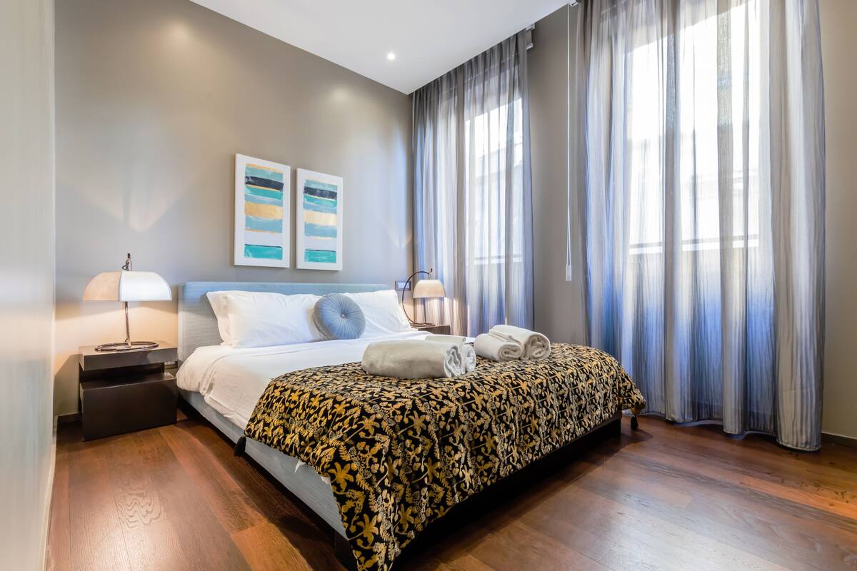 A modern hotel room with a large bed, two bedside tables with lamps, a view of the city through sheer curtains, and artwork on the wall.