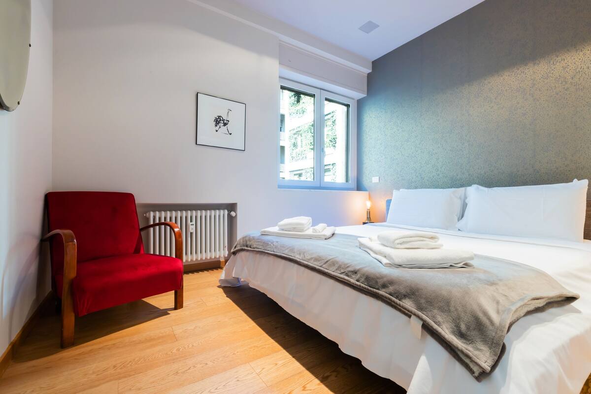 A hotel room with a bed, a red chair, a radiator, a window, and a framed picture on the wall.