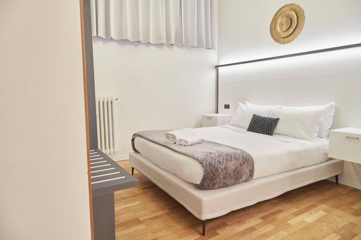 A modern bedroom with a bed, bedside tables, a radiator, and a wall-mounted decorative item.