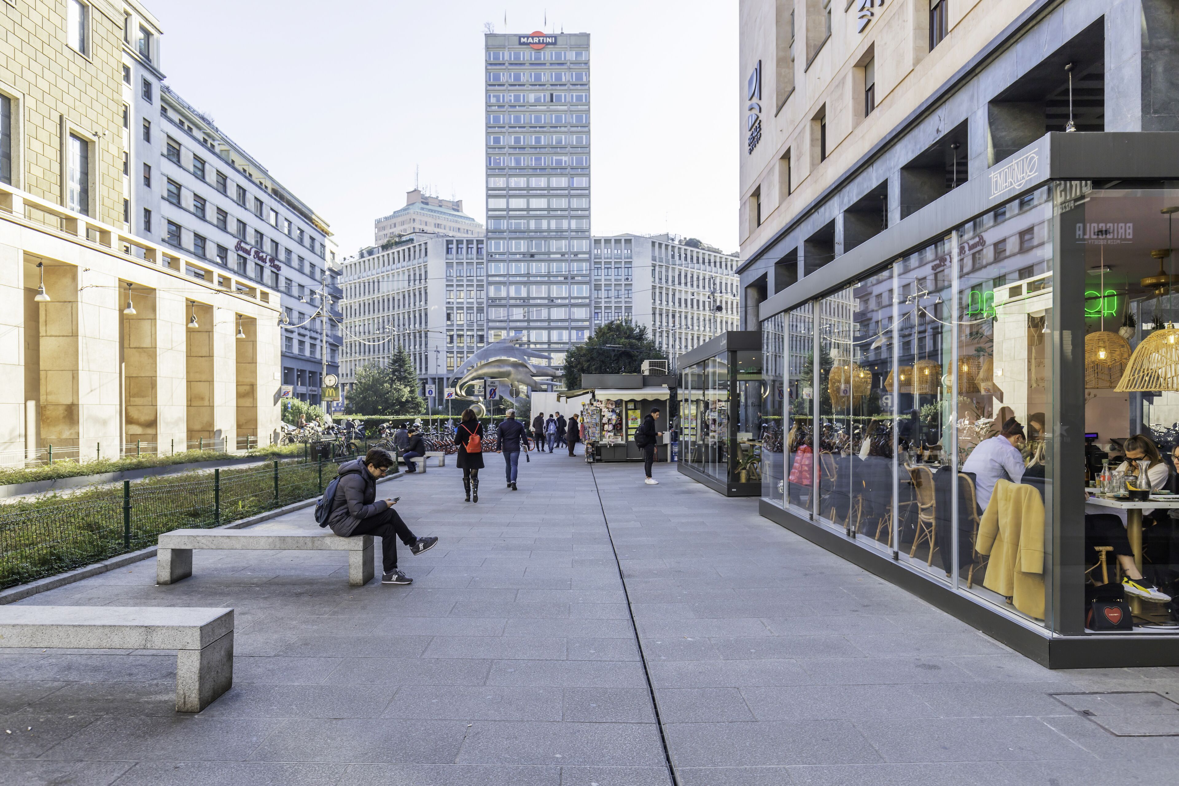 A pedestrian area with benches, shops, and a tall building in the background.