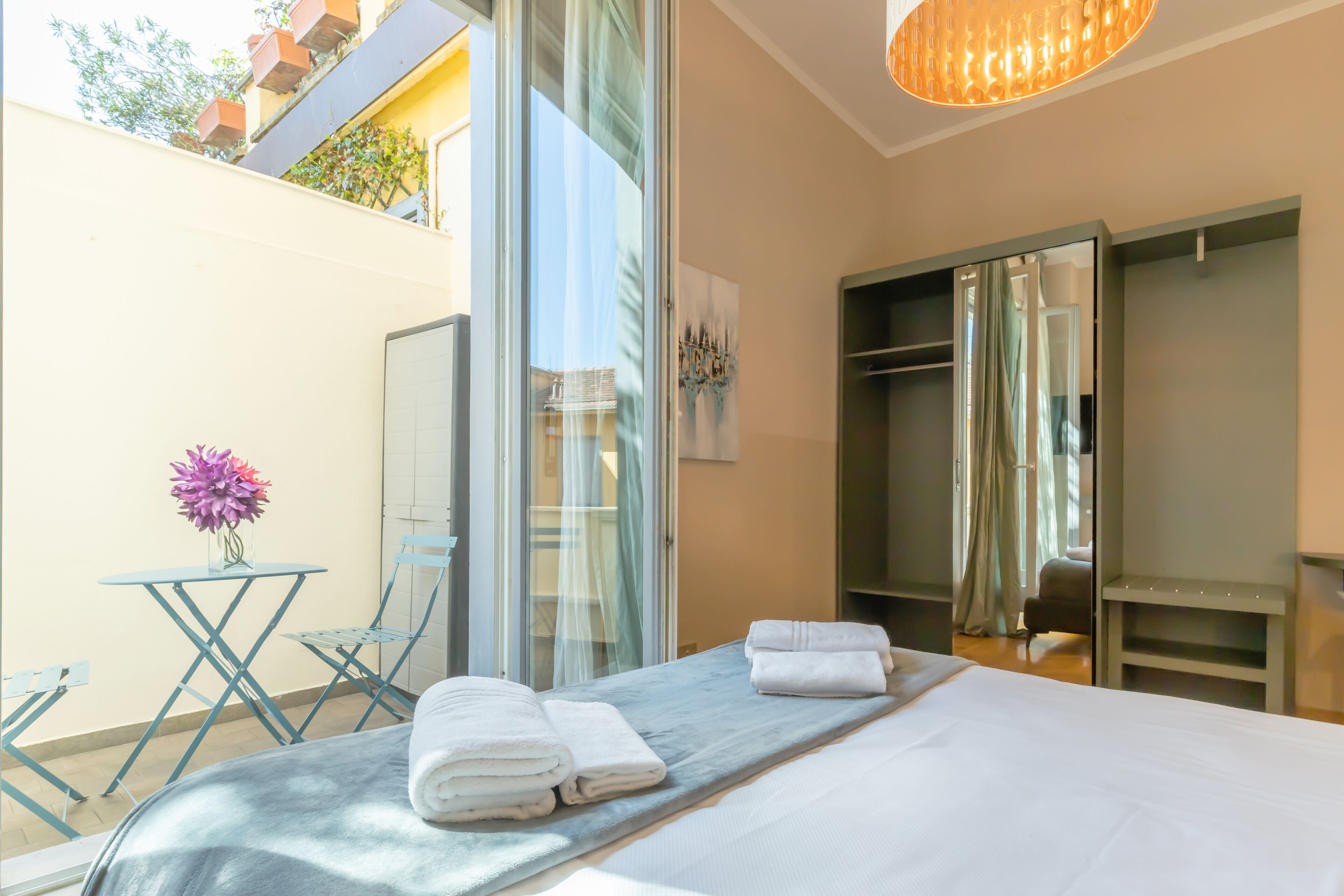 Een hotelkamer met een bed, een klein tafeltje met een bloemenvaas, een stoel en een glazen schuifdeur naar een balkon.