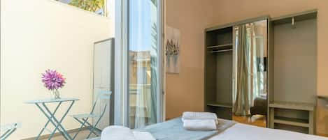 A hotel room with a bed, a small table with a vase of flowers, a chair, and a sliding glass door leading to a balcony.