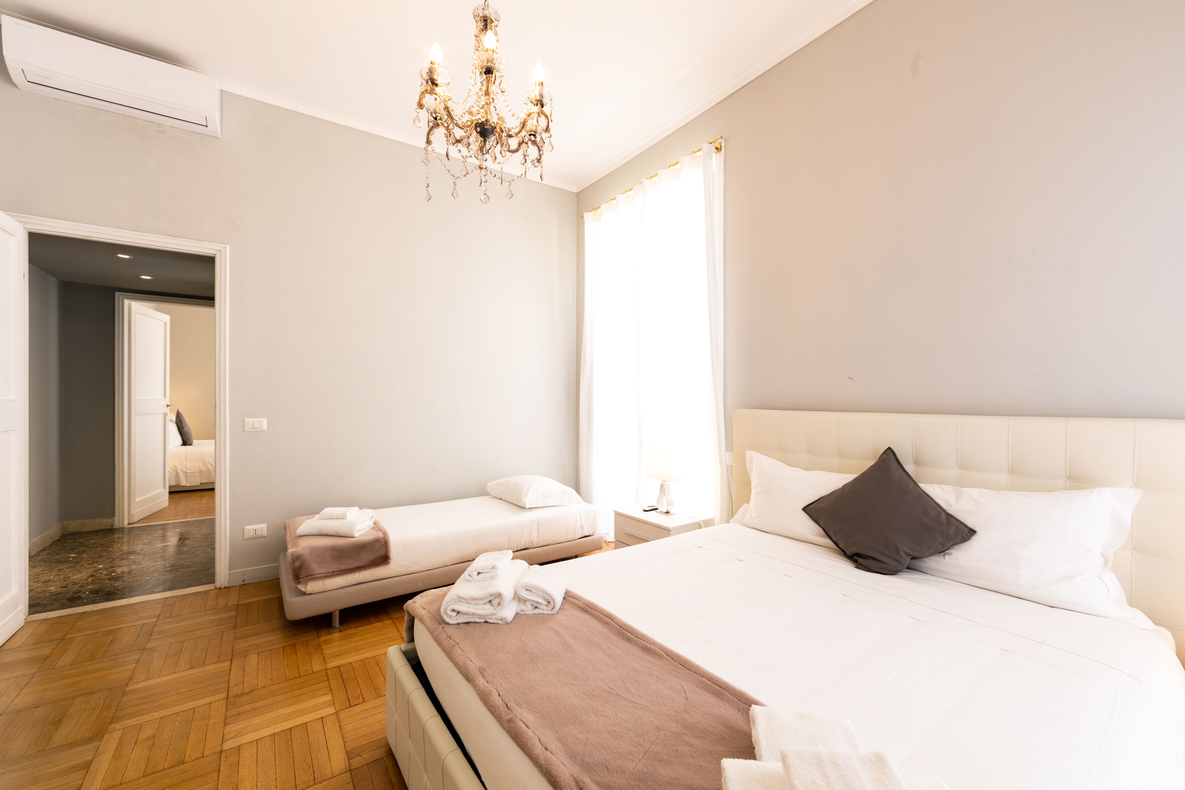 Hotelkamer met een groot bed, een kleiner bed, een kroonluchter, een raam en een deur naar een andere kamer.