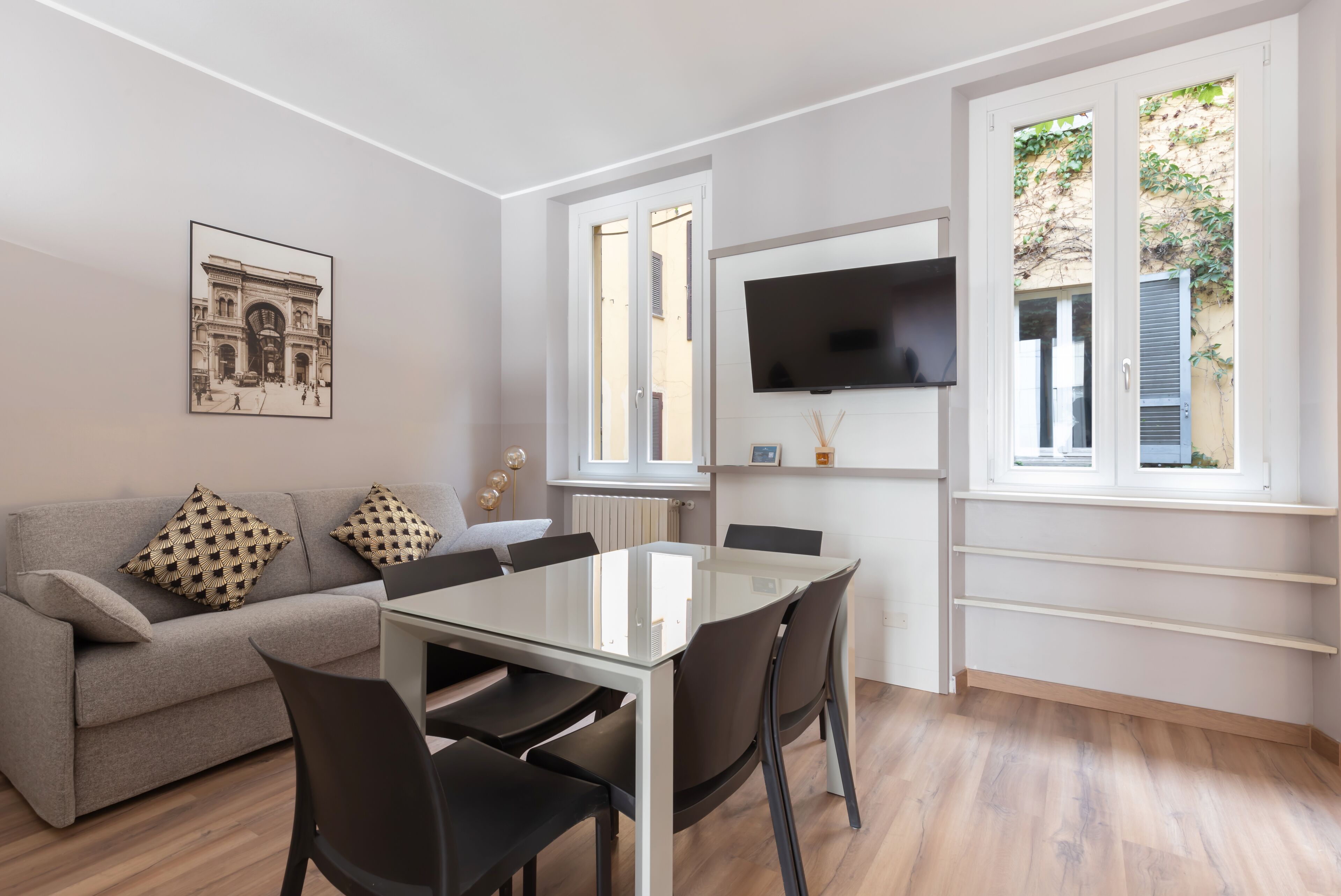A modern living room with a sofa, a glass-top dining table, and a TV mounted on the wall.