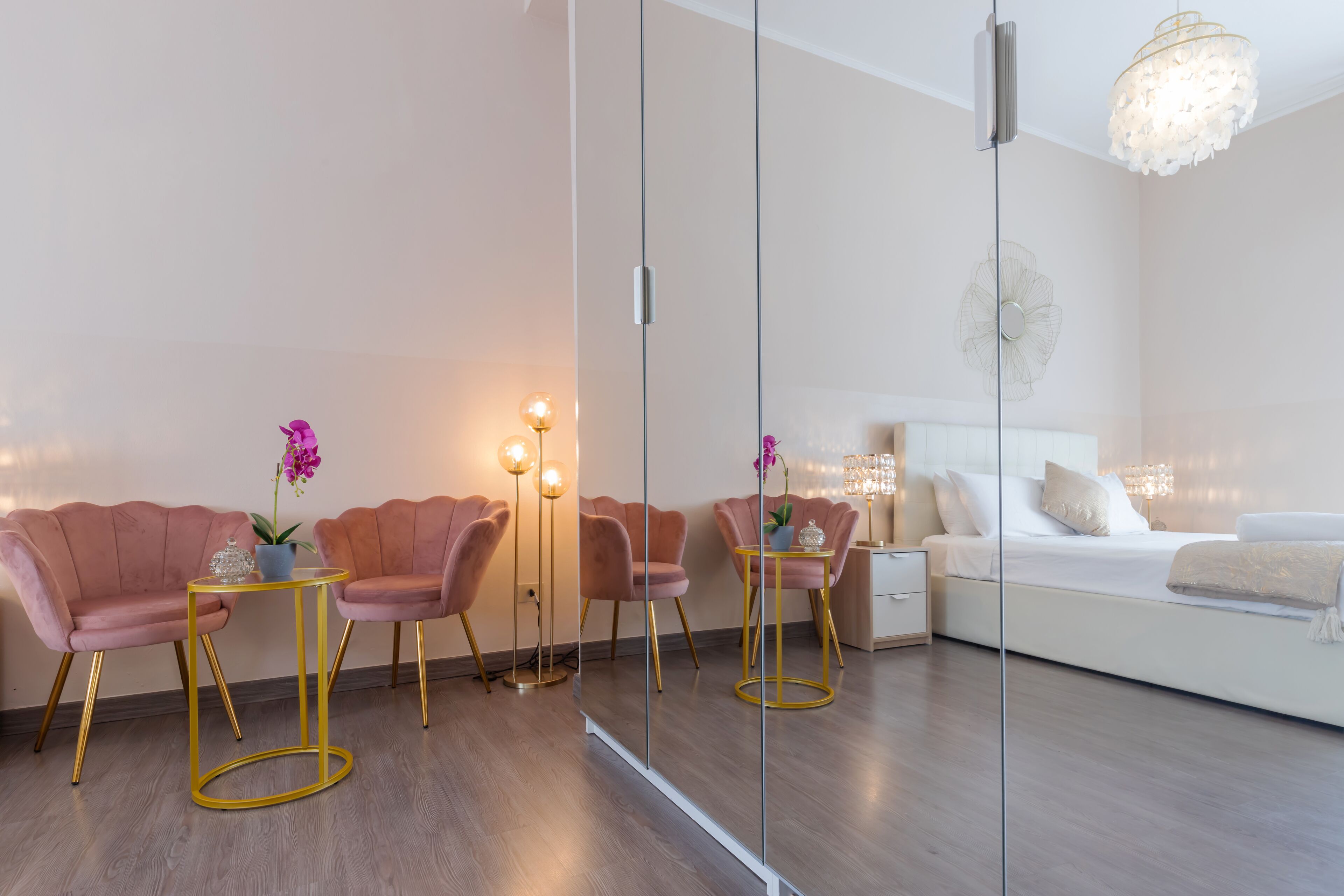 A modern bedroom with a large bed, bedside tables, and a mirror. There are two pink chairs with a small table and a floor lamp.