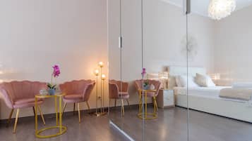 A modern bedroom with a large bed, bedside tables, and a mirror. There are two pink chairs with a small table and a floor lamp.
