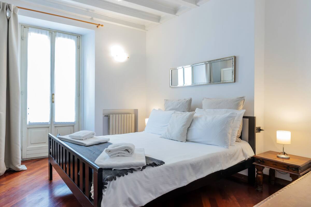 A neatly made bed with white linens, a bedside table with a lamp, and a large window with curtains.