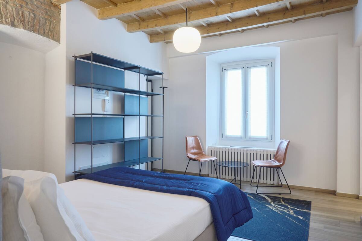 A bedroom with a bed, a blue bedspread, a bookshelf, two chairs, a window, and a radiator.