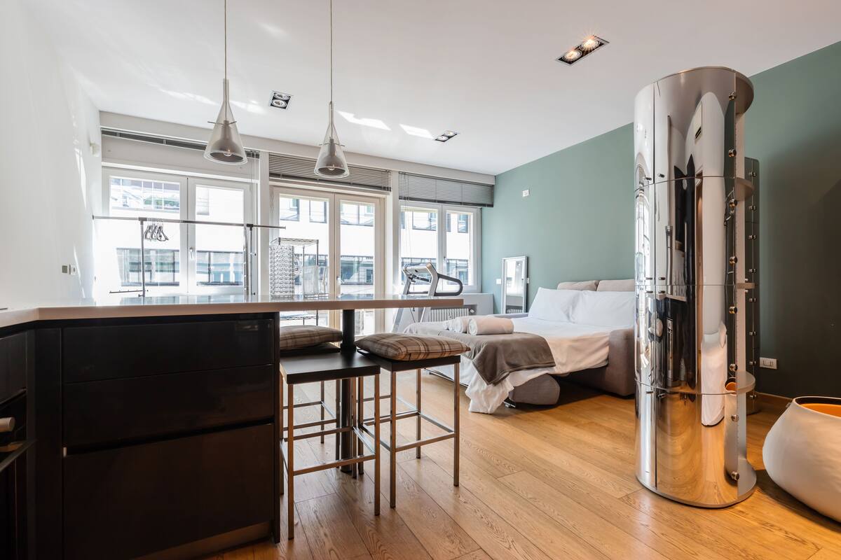 A modern hotel room with a kitchen area, a bed with a view, and a large cylindrical object that could be a decorative piece or appliance.