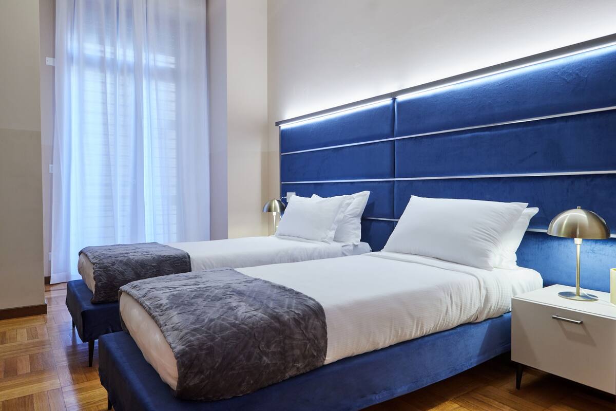 A hotel room with two beds, a blue headboard, white bedding, a bedside lamp, and a wooden floor.