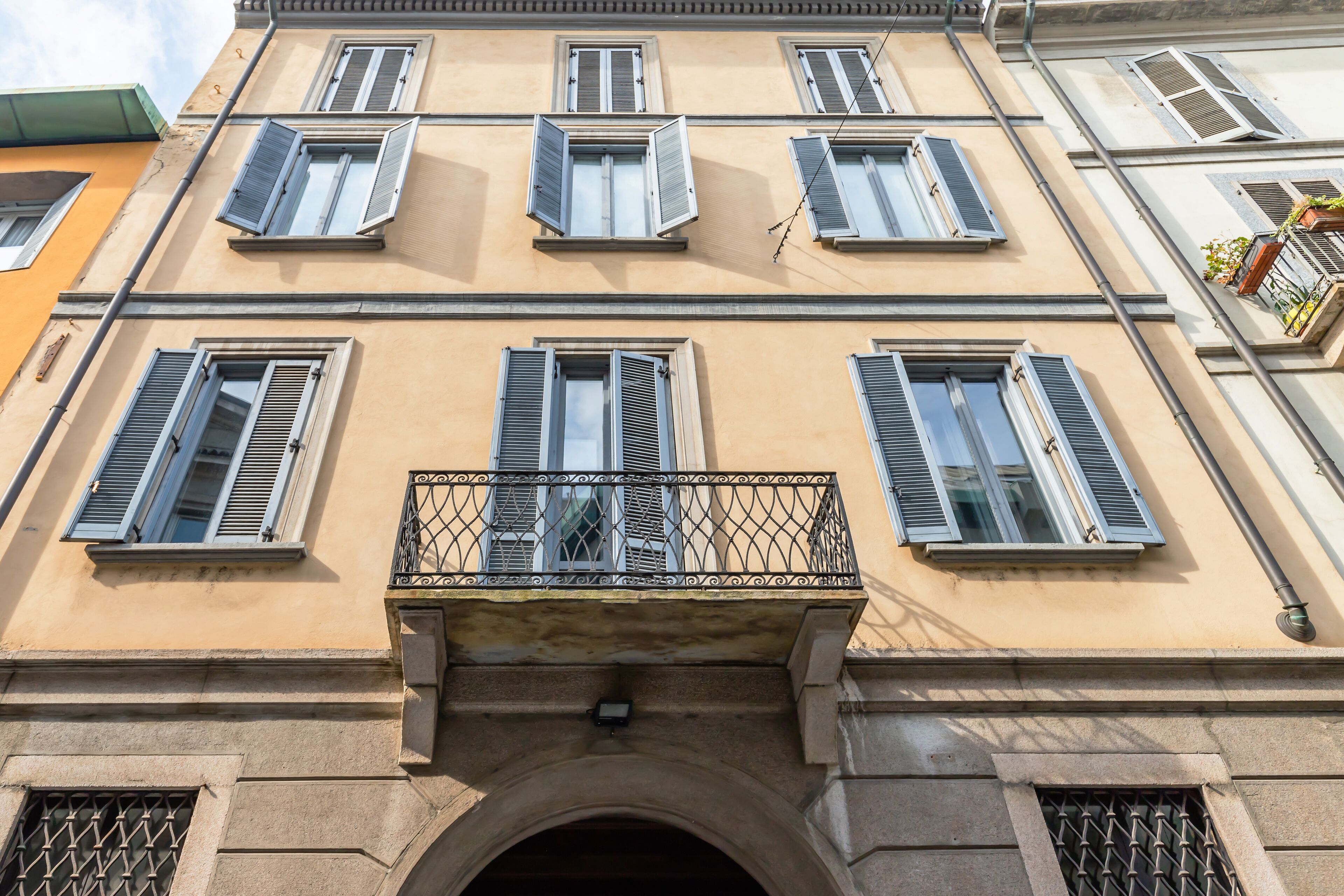 A multi-story building with a central balcony, large windows with shutters, and a stone archway at the entrance.