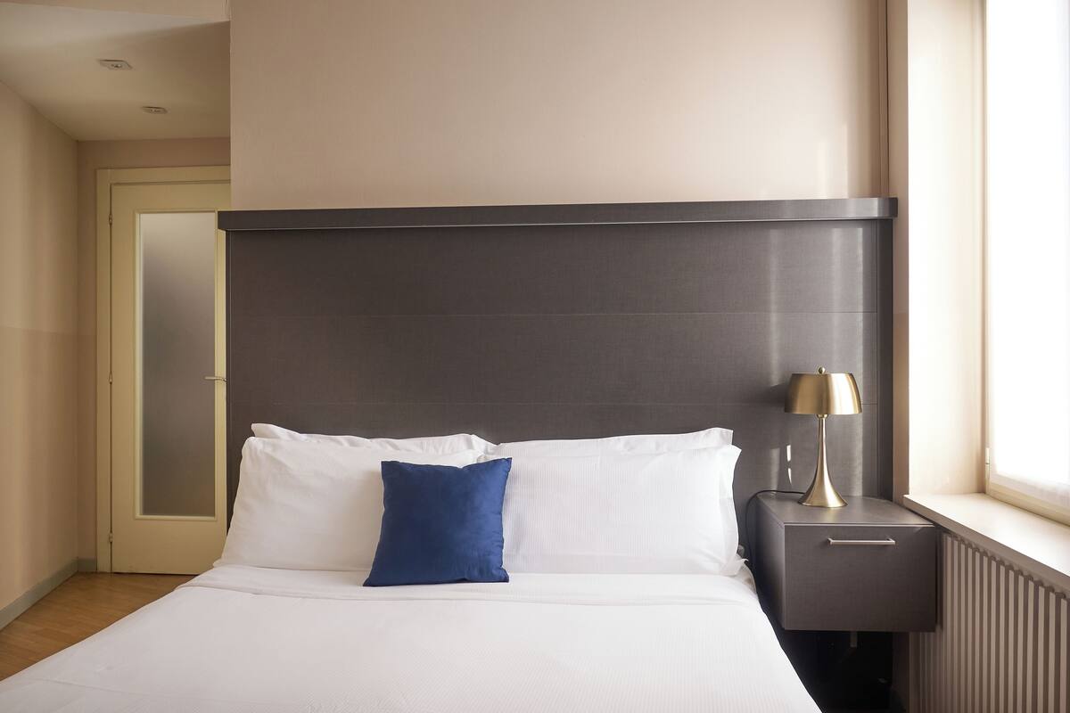 A modern hotel room with a large bed, a nightstand, a lamp, and a window.